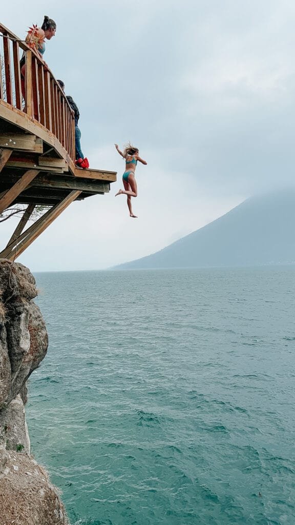 Woman in a bathing suit (an item that is at the top of a packing list for guatemala) jumping off the platform on Lake Atitlan with a volcano in the back